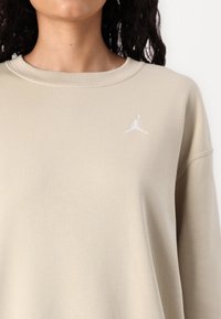 Béžová mikina s rebrovaným výstrihom a dlhými rukávmi. Obsahuje malé biele vyšité logo Jumpman na ľavej strane hrude.