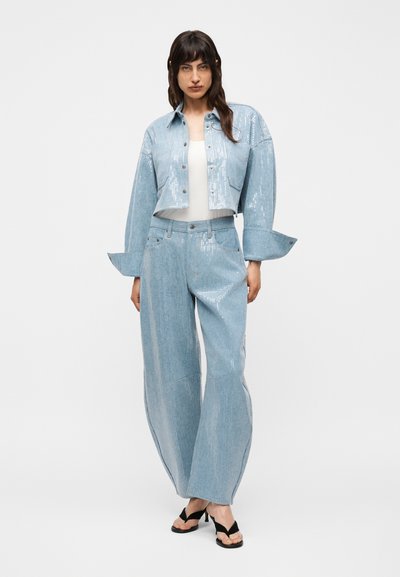 Γυναίκα με cropped σακάκι σε ανοιχτό γαλάζιο χρώμα με υφή παγιέτας και αντίστοιχη φαρδιά παντελόνια, λευκό τοπ από κάτω και μαύρα σανδάλια με τακούνι.