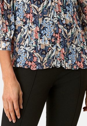 Kobieta w plisowanej bluzce w kwiatowy wzór w odcieniach niebieskiego, różowego i białego oraz w czarnych dopasowanych spodniach, z rękami swobodnie opuszczonymi wzdłuż ciała.