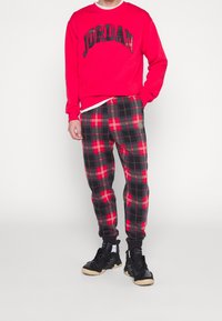 Rode sweatshirt met "JORDAN" in zwarte letters, gecombineerd met zwart-rode geruite joggers met een zijzak en zwarte sneakers.