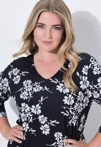 Junge Frau mit langen blonden Haaren, die ein schwarzes, blumenmusterndes V-Ausschnitt-Top trägt und mit den Händen in den Hüften vor einem weißen Hintergrund posiert.