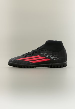 Zapato de fútbol negro con rayas rojas, suela con textura antideslizante y cuello alto similar a un calcetín, sobre un fondo liso.