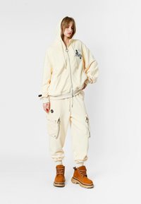 Joven mujer vestida con un sudadero crema y joggers de cargo a juego, con botas de cordones en tono beige, de pie frente a un fondo blanco.