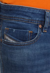 Vaqueros de mezclilla azul con un diseño clásico, que incluyen un bolsillo para monedas, detalles de costura en naranja y un cierre de botón metálico.
