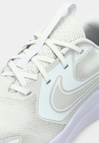 Zapatilla deportiva de color gris claro con parte superior de malla, detalles sintéticos lisos, cordones planos y suela blanca con un sutil detalle de logo.