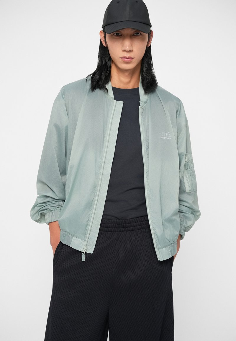 Giacca a bomber leggera di colore verde chiaro, con zip frontale, polsini elastici e una tasca sul maniche. Indossata sopra una maglietta scura e pantaloni corti neri.