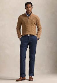 Mann står med hendene i lommene, iført en beige kabelstrikket genser, marineblå bukser, brunt belte og brune skinnsko.