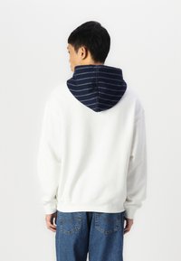 Biela mikina s kapucňou s námorníckymi pruhmi. Tkanina vyzerá mäkko, má voľný strih a ribbed manžety. V kombinácii s modrými džínami.