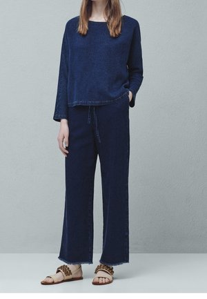 Mørkeblå, struktureret strik bluse med lange ærmer og rund halsudskæring, sat sammen med vide, marineblå bukser. Lyse sandaler med blomstrede detaljer.