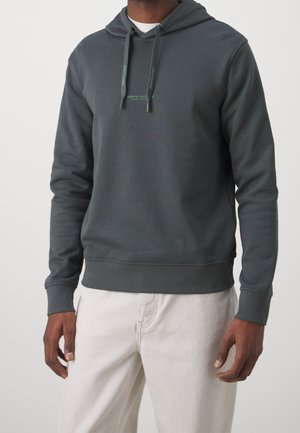 Man draagt een donkergrijze hoodie met trekkoorden en witte tekst op de borst, gecombineerd met lichtbeige broek tegen een effen achtergrond.