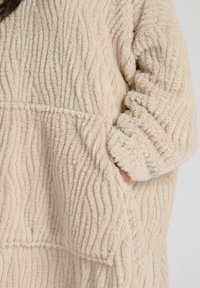 Pullover plush beige con texture, dal morbido superficie a coste, caratterizzato da una vestibilità ampia e tasche laterali per maggiore funzionalità.