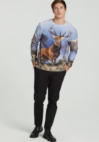 Sudadera gráfica de manga larga que presenta un estampado de ciervo en estilo acuarela en tonos azules, marrones y verdes. Pantalones y zapatos negros completan el atuendo.