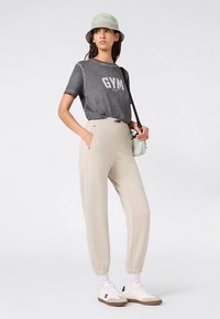 T-shirt gris avec inscription "GYM", pantalon beige avec poignets élastiques, baskets blanches et bob vert clair. Sac épaule minimaliste.