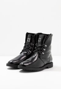 Botas de tobillo de cuero negro con acabado brillante, con diseño de cordones, forro de piel sintética y suela de goma texturizada. Incluye detalles de hardware.