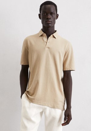 Beige short-sleeved polo shirt with a textured fabric, buttoned collar, relaxed fit, and side seam detail. Paired with white linen trousers.