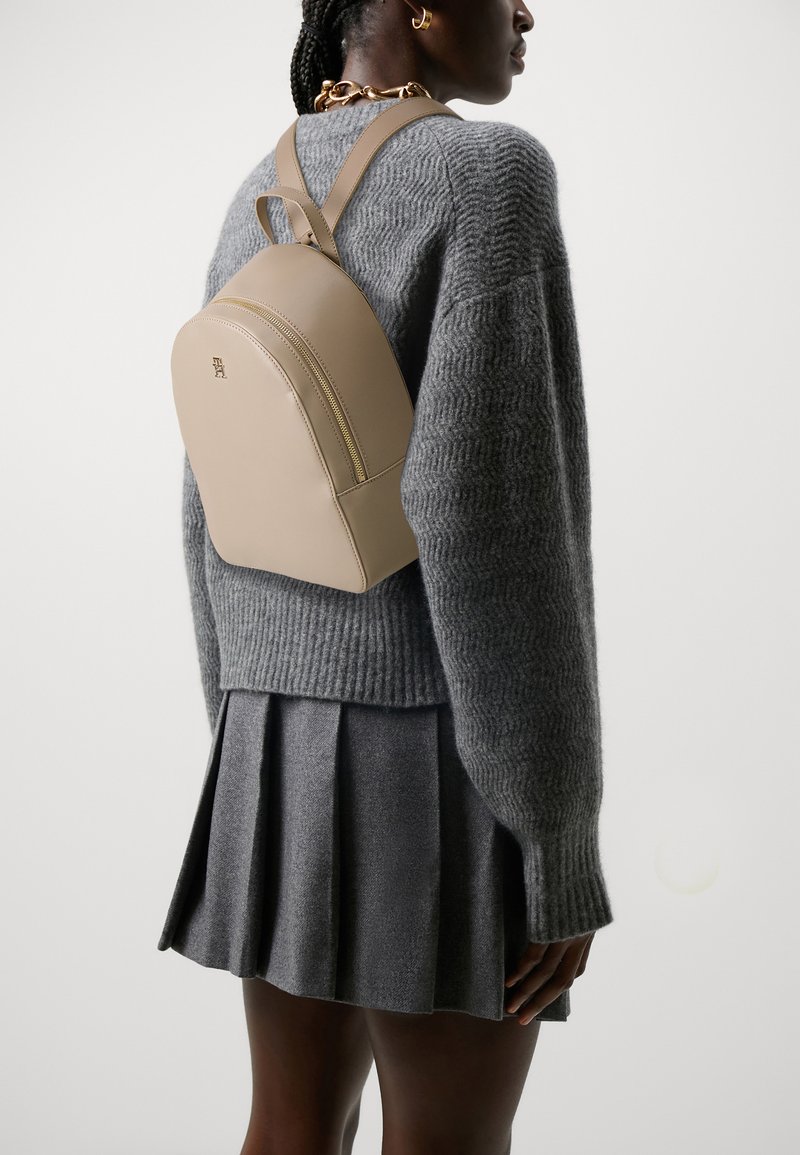 Zaino in pelle beige dalla forma arrotondata, con una cerniera frontale e spallacci regolabili, indossato sopra un maglione grigio a maglia e una gonna grigia.