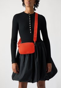 Borsa a tracolla in pelle rossa con texture, chiusura a zip liscia e tracolla regolabile, indossata sopra un top nero a coste e una gonna nera voluminosa.