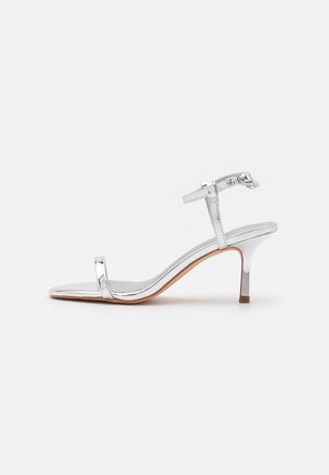 Silberne High-Heel-Sandale mit dünnem Vorderriemen, Knöchelriemen mit Schnalle und einem schlanken mittelhohen Absatz auf hellem Hintergrund.