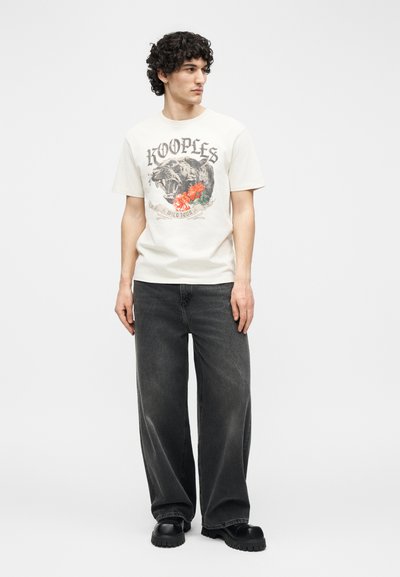 Νέος άνδρας φοράει λευκό μπλουζάκι με γραφικό απεικόνιση ένα λιοντάρι που βρυχάται και κόκκινα λουλούδια, χαλαρά μαύρα τζιν και μαύρα χοντρά παπούτσια, στέκεται και κοιτάζει προς τα αριστερά.