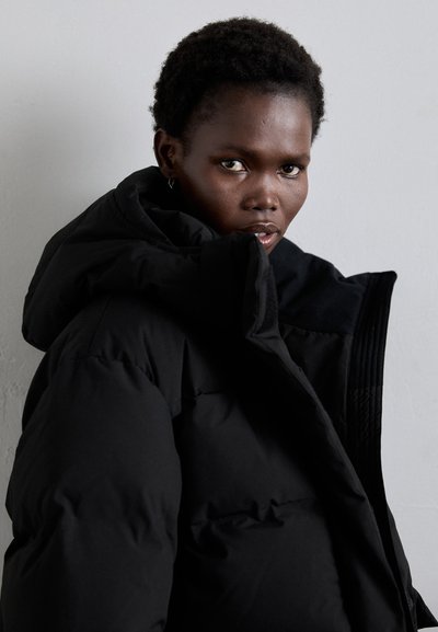 Black padded jacket featuring a large hood, zippered front, and textured accents on the shoulders. Smooth fabric and oversized fit.