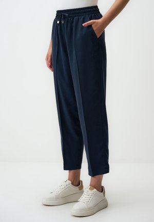 Persoon die marineblauwe taps toelopende broek draagt met elastische tailleband en witte sneakers met beige accenten, staand tegen een witte achtergrond.