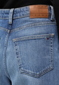 Πίσω τσέπη από μπλε τζιν με μικρό λογότυπο Tommy Hilfiger και καφέ δερμάτινη ετικέτα με την ένδειξη Tommy Jeans στη μέση.