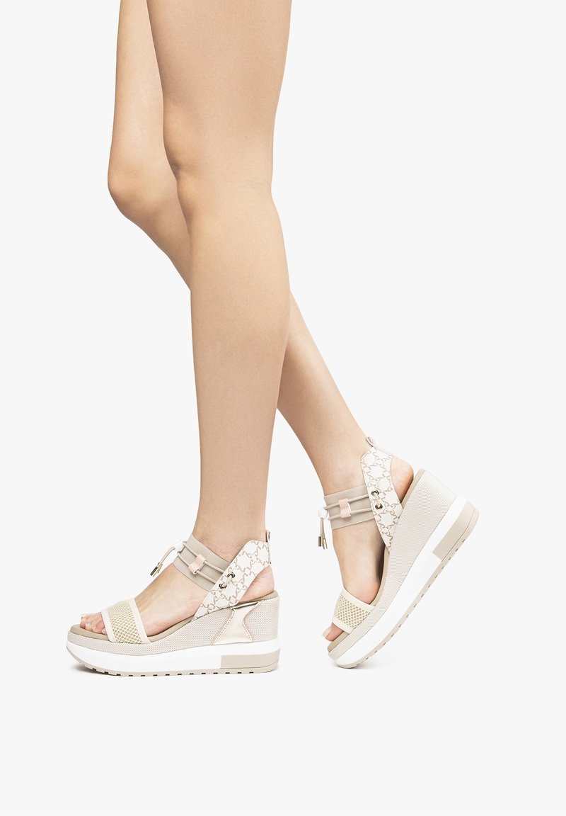 Jambes portant des sandales compensées beige avec des larges brides, des orteils ouverts et des brides à motif aux chevilles, debout sur fond blanc.
