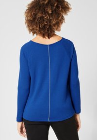 Blå långärmad topp med rund halsringning och en bakre söm med vit stickning. Tillverkad av mjukt, texturerat tyg, figursydd vid midjan.