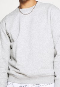 Sudadera de cuello redondo gris claro, hecha de una tela suave con un ajuste clásico, que presenta puños y dobladillo acanalados, complementada con un collar de cadena plateada.
