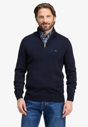 Homme portant un pull bleu marine à demi-fermeture éclair par-dessus une chemise fleurie bleue avec un jean bleu, debout devant un fond blanc uni.