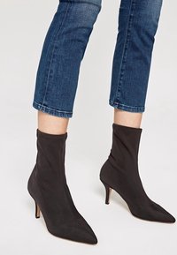 Úzké černé kotníkové boty s špičatou špičkou a podpatky typu kitten, nošené se zkrácenými modrými džínami na světlém pozadí.