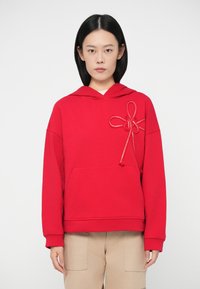 Γυναίκα που φοράει μια κόκκινη φούτερ με διακοσμητικό κορδόνι στο στήθος και μπεζ παντελόνι, στέκεται μπροστά σε λευκό φόντο.