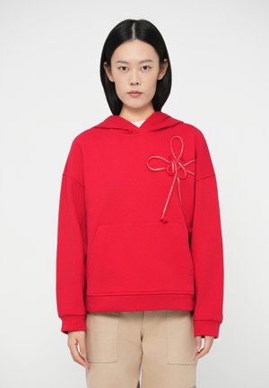 Frau in einem roten Hoodie mit einer dekorativen Seil-Schleife auf der Brust und beigen Hosen, die vor einem weißen Hintergrund steht.