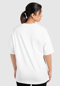 Camiseta de algodón blanca con un corte holgado, mangas cortas y cuello redondo, vista desde atrás sobre un fondo gris claro.