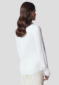 Biała, długa, lniana koszula z klasycznym kołnierzykiem, z zapięciem na guziki przy mankietach oraz zaokrąglonym dołem. Lekki, oddychający materiał o gładkiej strukturze.