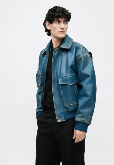 Νεαρός άνδρας που φοράει ένα φθαρμένο μπλε δερμάτινο μπουφάν πάνω από μαύρο πουκάμισο και παντελόνι, στέκεται με τα χέρια στις τσέπες κοιτάζοντας προς τα πλάγια μπροστά σε λευκό φόντο.