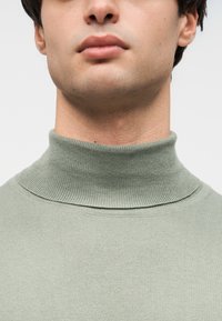 Πλεκτό πράσινο πουλόβερ με ριγωτή υφή και ψηλό λαιμό, με εφαρμοστή γραμμή και φαρδιά ρίγα στον γιακά. Χωρίς ορατά σχέδια ή διακοσμητικά στοιχεία.