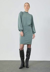Um vestido verde-água com mangas longas e bufantes e uma saia ajustada, feito de um tecido suave, combinado com botas de couro pretas até ao joelho.