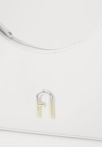 Sac à main en cuir blanc avec un design incurvé, présentant un accent en métal argenté et doré sous la forme d'une lettre stylisée. Fermeture zippée.