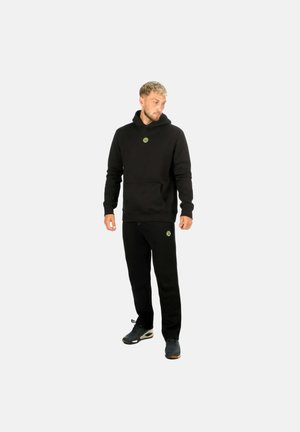 Mann trägt schwarzen Hoodie und Jogginghose mit kleinen grünen kreisförmigen Logos, steht und schaut nach unten links.