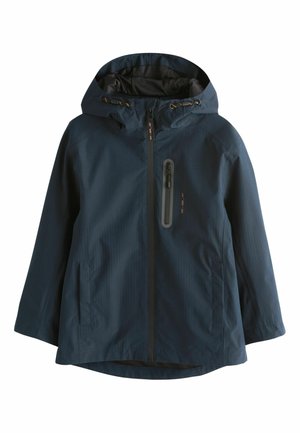 Person iført en mørk marineblå jakke med lodret lynlåslomme på brystet og trykte koordinater på ærmet, arme krydset over brystet.