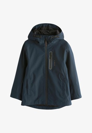 Person iført en mørk marineblå jakke med lodret lynlåslomme på brystet og trykte koordinater på ærmet, arme krydset over brystet.