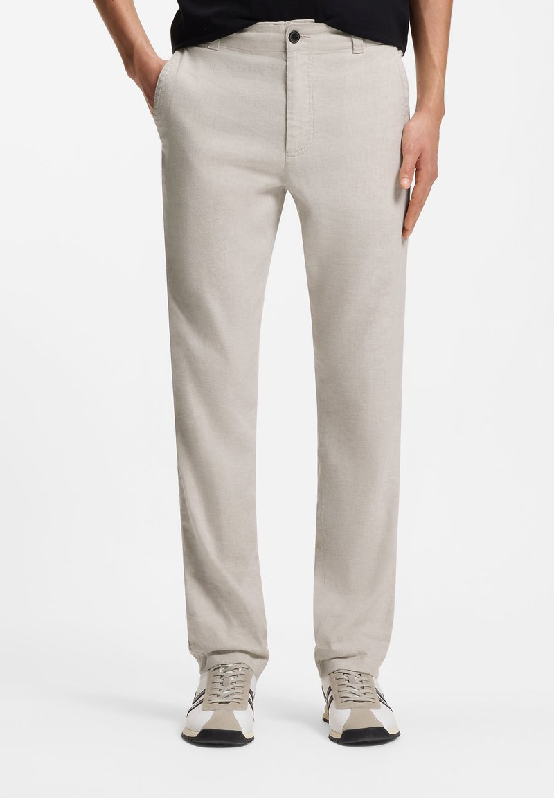 Pantalones beige claro ajustados combinados con zapatillas casuales blancas y beige, manos en los bolsillos, mostrados desde la cintura hasta los pies sobre fondo blanco.