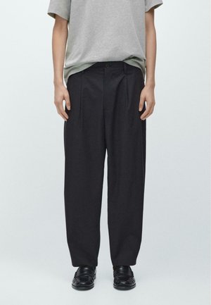 Osoba nosząca luźne czarne plisowane spodnie, szarą koszulę z krótkim rękawem i czarne loafersy, stojąca na tle jednolitego jasnego tła.