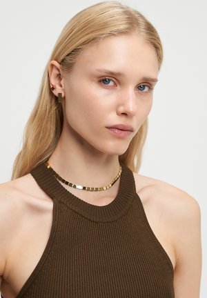 En blond kvinde med blå øjne bærer en brun ribbet halterneck-top, et kvadratisk guldhalskæde og matchende kvadratiske guldringe foran en ensfarvet baggrund.