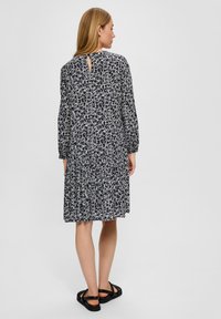 Schwarzes Kleid mit einem weißen floralen Muster, langen Ärmeln, gestuftem Design und einem Schlüssellochöffnung im Rücken. Kombiniert mit schwarzen, riemenartigen Sandalen.