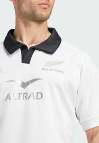 Hvid polo-shirt med sort krave, prydet med sølv logoer for ALTRAD og All Blacks. Kortærmet med en glat tekstur og sporty design.