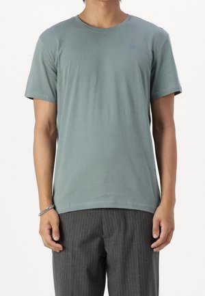 Mann, der ein einfaches, gedämpft grünes kurzärmeliges T-Shirt und dunkelgraue Nadelstreifenhose trägt, steht mit entspannten Armen.