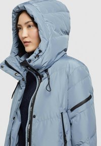 Donna che indossa una giacca invernale imbottita di colore blu chiaro con un ampio cappuccio e dettagli neri sulla cerniera, che guarda di lato su uno sfondo neutro.