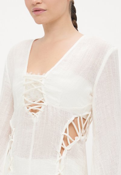 Vêtement blanc texturé présentant un décolleté en V profond, des détails à lacets et des côtés découpés. Les manches longues avec des accents élastiques ajoutent un détail de design.